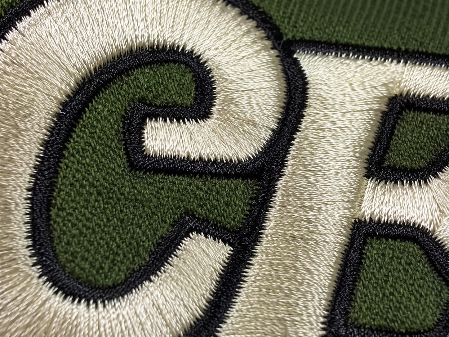 Macro close-up of embroidered green and cream block letters on a dark green garment, showing the textured stitching of each character